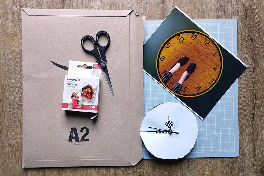Crafting materials including scissors, photo tape, a clock face, and an a2 cardboard sheet on a wooden surface.