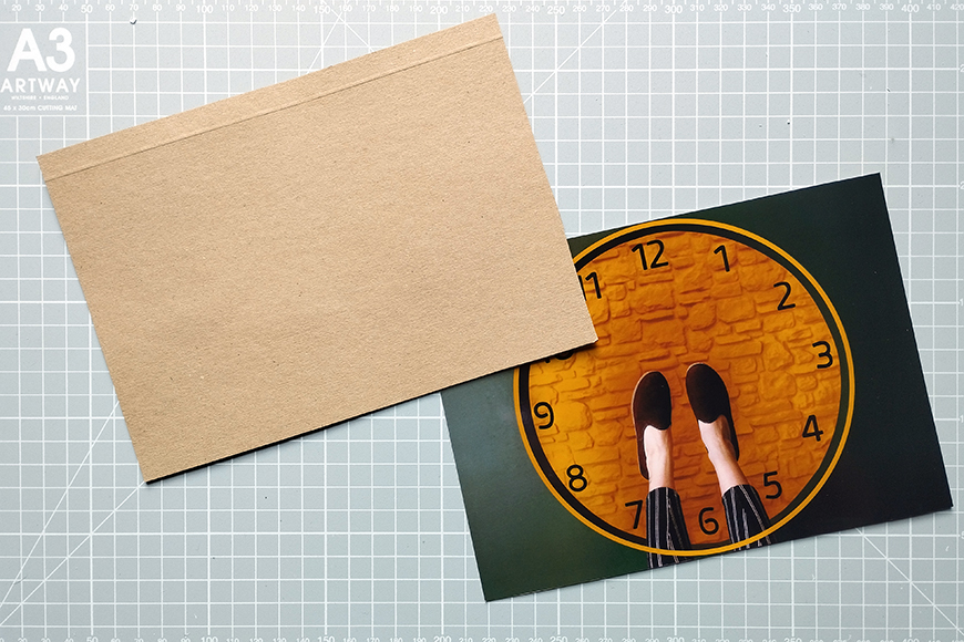 A brown envelope partially covering a greeting card with a quirky design of shoes on a clock face, resting on a cutting mat.