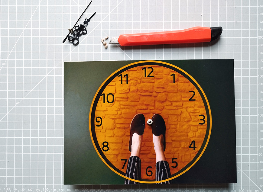 Art and craft station with printed clock hands on a brick background, small figure, scissors, and cutter knife on a cutting mat.