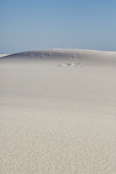 White sand dunes under a clear blue sky, with subtle wind patterns on the sand's surface.