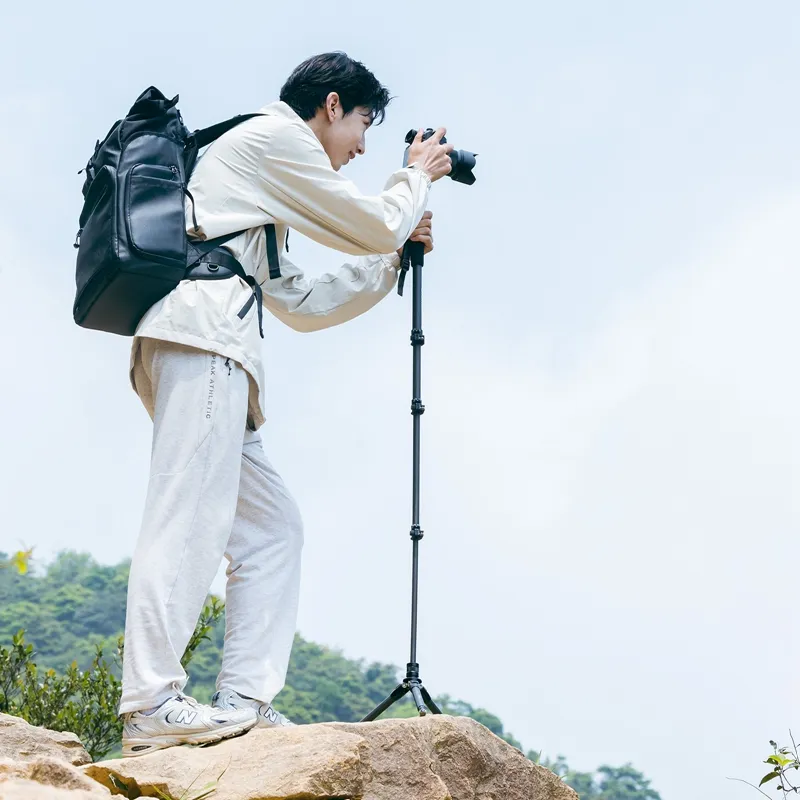 A man with a backpack using a camera mounted on a tripod atop a rocky outlook with lush hills in the background.