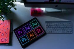 A desktop with an imac, a keyboard, a graphics tablet displaying adobe software icons, and decorative items including a plant and a neon pink paperweight.