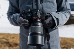 Person holding a sony camera with a zoom lens, wearing gloves, with a snowy field in the background.