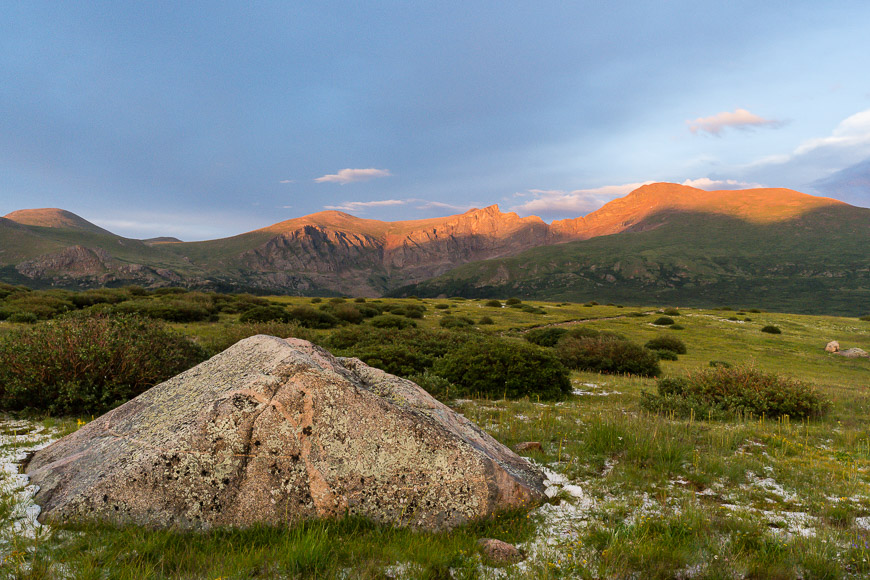 Alpenglow illuminates mountain peaks during sunset with a large rock in the foreground.
