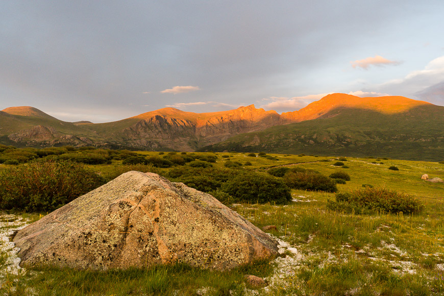 Alpenglow on mountain peaks at sunset with a prominent boulder in the foreground.