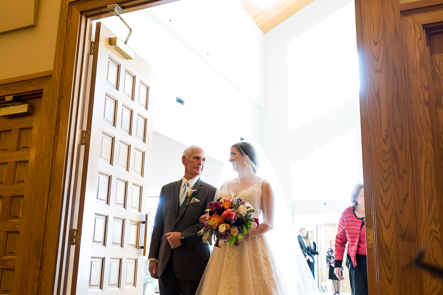 A bride accompanied by an older gentleman walking down the aisle, bathed in natural light from a large window.