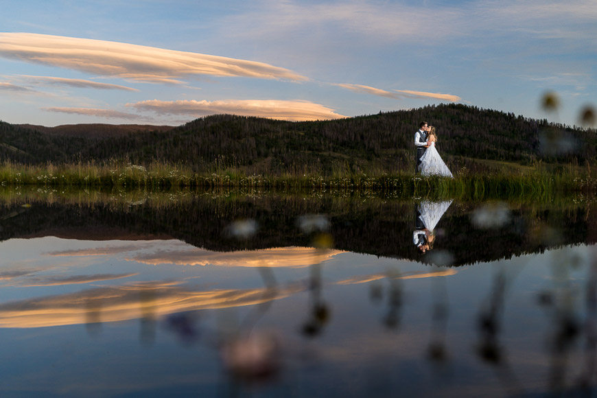 Couple embracing on lakeshore with reflection and sunset.