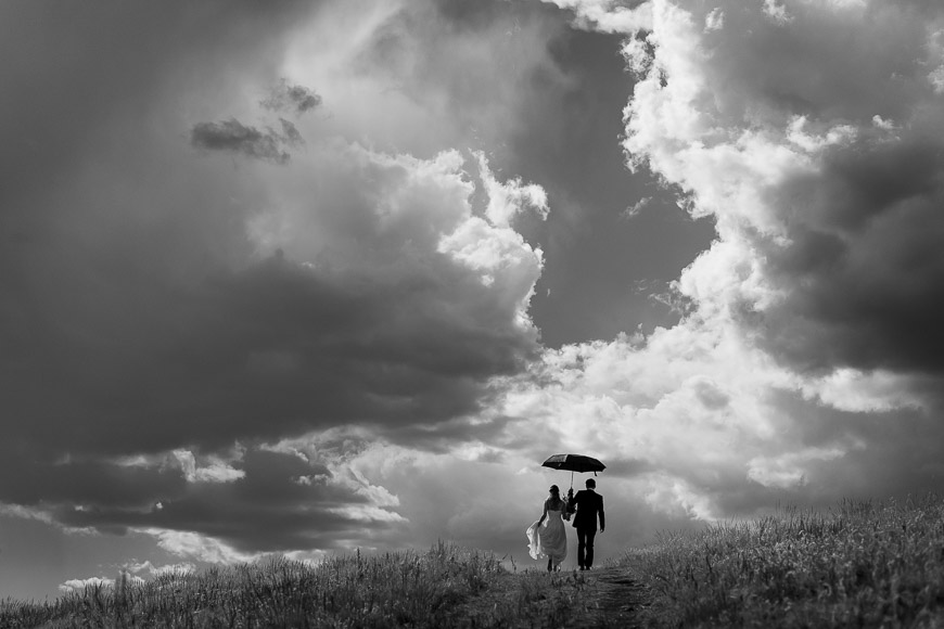 Two people walking under an umbrella on a path with a dramatic cloudy sky above.