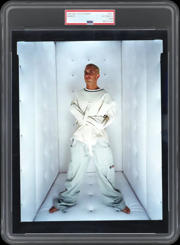 A person in a white outfit posing inside a confined, illuminated space, resembling an artistic installation.