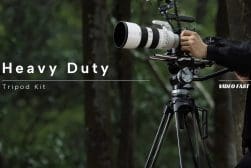 Hands adjusting a large camera mounted on a heavy-duty tripod in a forest setting, with text overlay reading "heavy duty tripod kit.