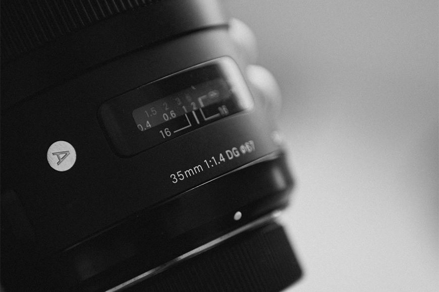 Close-up black and white photo of a camera lens with focus distance and aperture markings visible.