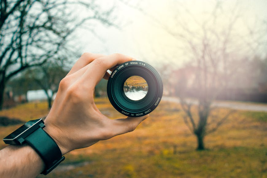A hand holding a camera lens which captures a reversed image of a tree-lined landscape, emphasizing the lens's clarity and focus.