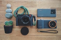 Top view of a neatly arranged leica camera, smartphone, portable speaker, pile of stones, and a small potted plant on a wooden surface.