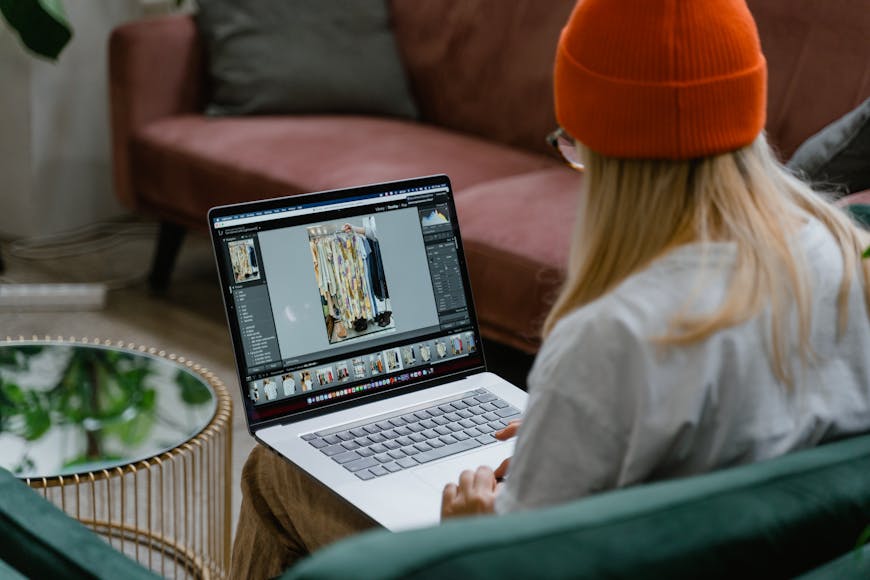 A woman in a beige sweater and orange beanie using a laptop on a couch, editing a fashion photo.