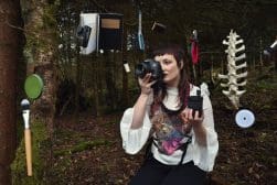A woman with a camera in a forest surrounded by various items like books and utensils hanging from trees.