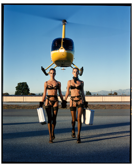A helicopter hovering above two models carrying briefcases on a runway.