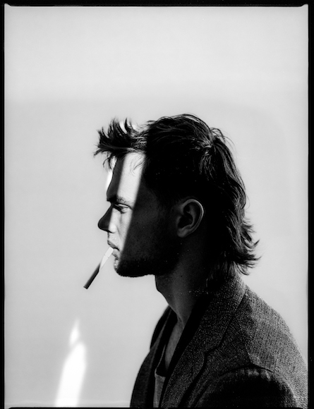 Profile of a man with tousled hair and a cigarette in his mouth, against a light background.
