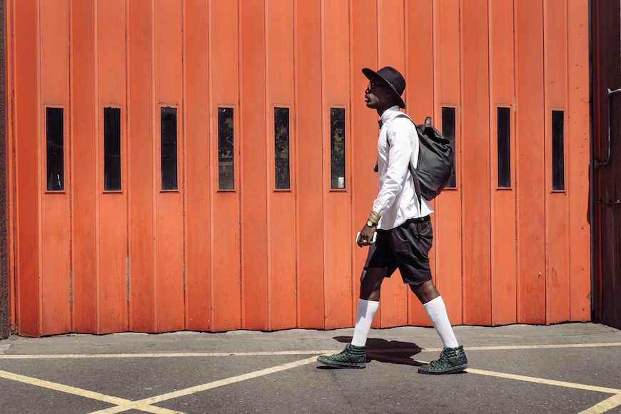 A man walking with a backpack in front of an orange building.