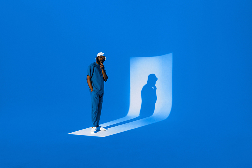 A man in blue clothing and a white hat stands on a white rectangle in a blue room, casting a shadow on a curved white backdrop.