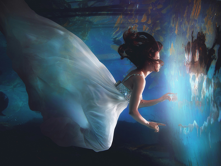 A woman in a white dress floats underwater, reaching toward a glowing, reflective surface in front of her.