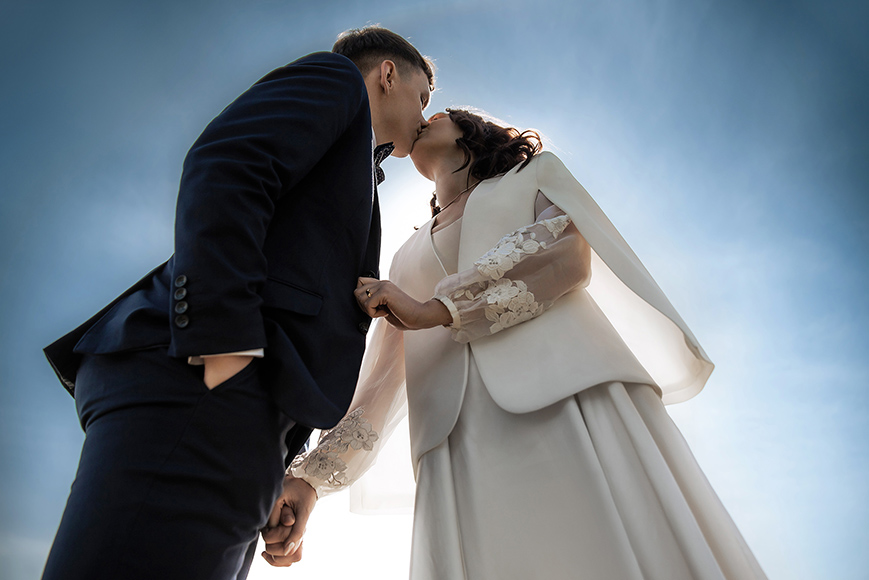 A couple dressed in formal attire hold hands and share a kiss outdoors under a clear blue sky, seen from a low angle.