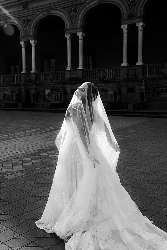 A bride in a long, flowing wedding dress and veil stands outdoors, tilting her head back, with an ornate building in the background.