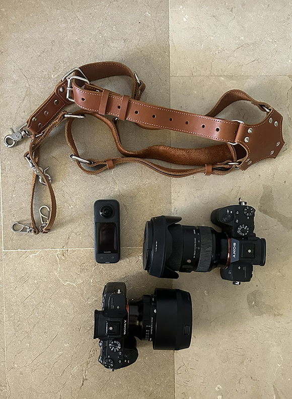 A brown leather camera harness, two Sony cameras with attached lenses, and a compact black 360-degree camera on a tiled floor.