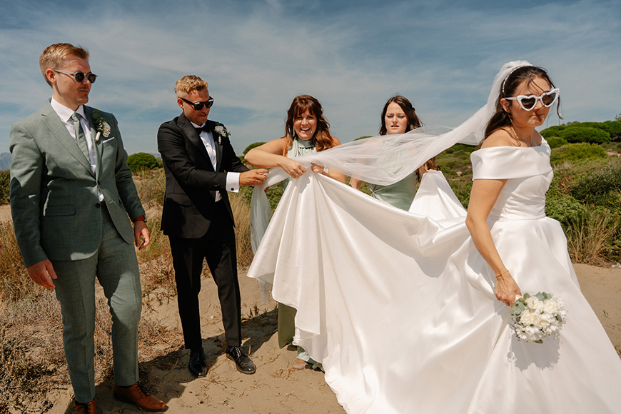 A bride in a white dress and sunglasses stands outdoors, while her veil and train are adjusted by three attendants and a man in a suit. Another man in a green suit stands nearby.