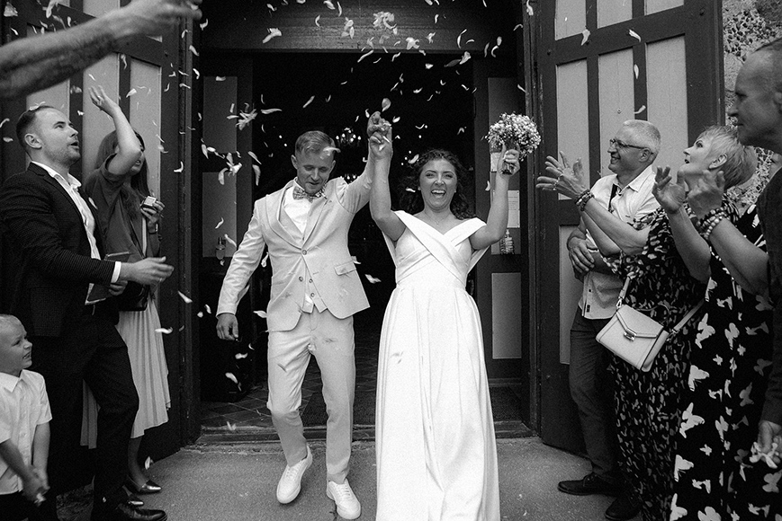 A newlywed couple exits a building holding hands while guests on both sides throw confetti to celebrate their marriage.