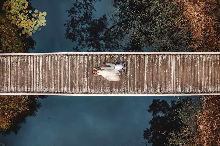 A bride and groom lie on a wooden bridge over water, surrounded by greenery, viewed from above.