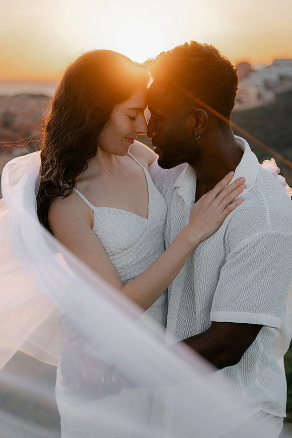 A couple in white clothing embraces outdoors at sunset, touching foreheads and surrounded by flowing fabric.