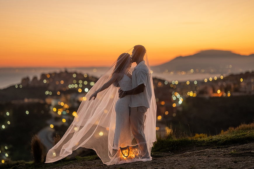 A couple in wedding attire stands outdoors at sunset, embracing under a sheer veil with city lights blurred in the background.