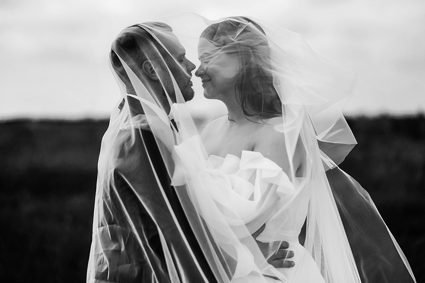 A bride and groom embrace outdoors, smiling at each other under a shared veil, with a blurred natural background.