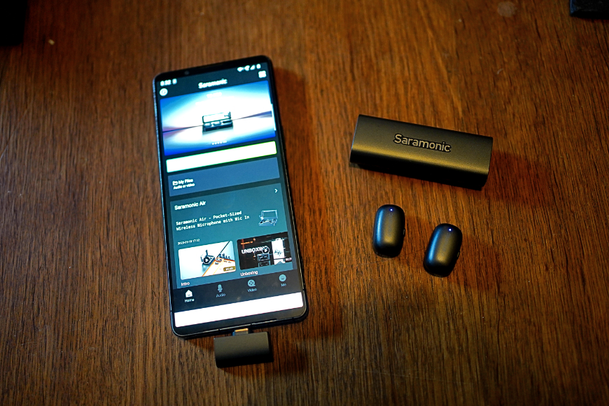 A smartphone with an audio app open sits on a wooden surface next to a Saramonic earbud charging case and two wireless earbuds.