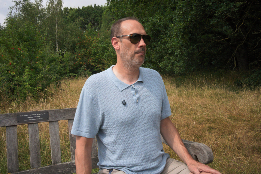 A man wearing sunglasses and a light blue polo shirt sits on a wooden bench outdoors, with tall grass and trees in the background.