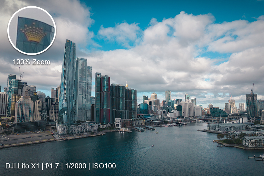 A city skyline with high-rise buildings along a waterfront under a partly cloudy sky; zoomed detail of one building is shown in an inset. Camera specs are displayed at the bottom.