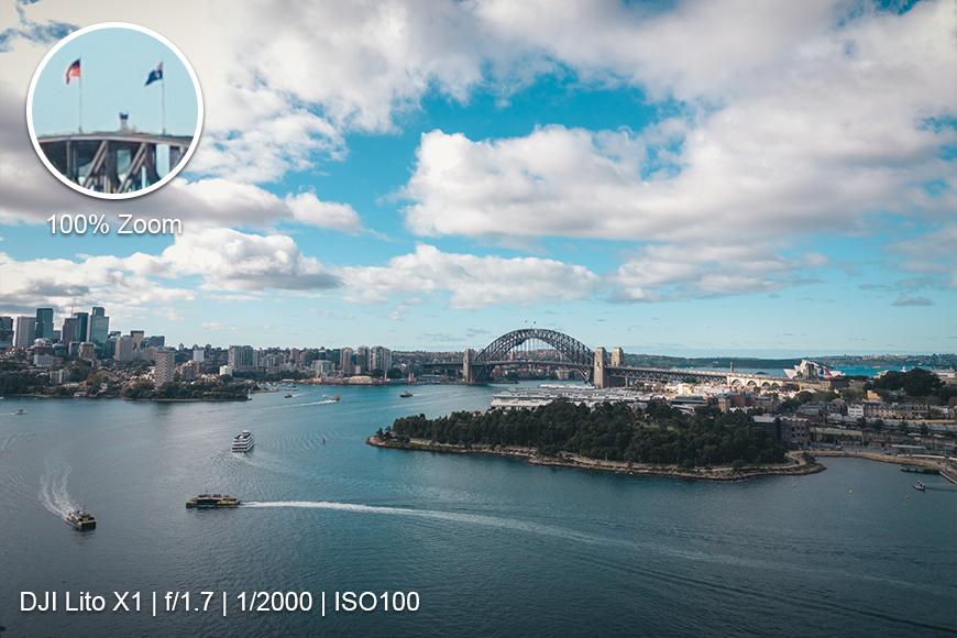 Aerial view of Sydney Harbour with the Sydney Harbour Bridge, city skyline, boats on the water, and camera settings overlaid in the bottom left corner. Inset shows a zoomed-in detail of bridge flags.