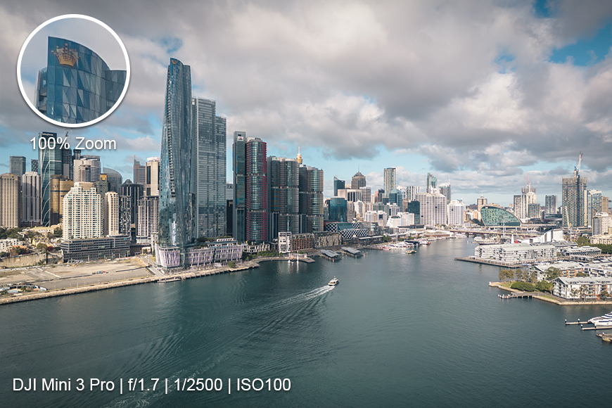 Aerial view of a modern city skyline with high-rise buildings along a waterfront. Text overlay shows camera settings and a zoomed-in detail of one building.