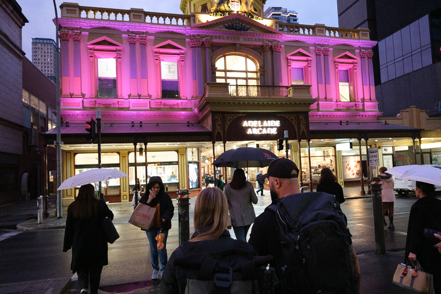 People with umbrellas cross a wet street toward a brightly lit Adelaide Arcade building at dusk, illuminated in pink and gold lights.