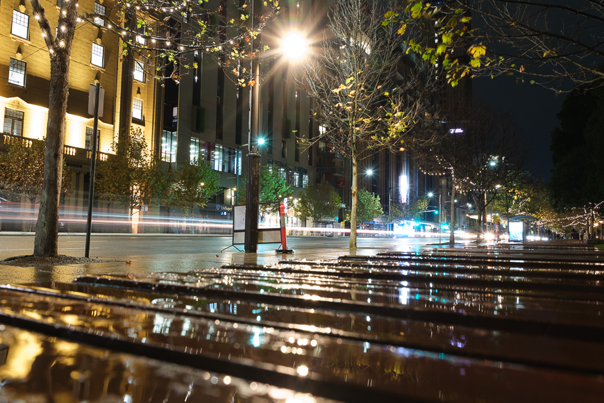 City street at night with wet benches in the foreground, illuminated by streetlights, passing light trails from vehicles, and buildings with lit windows in the background.