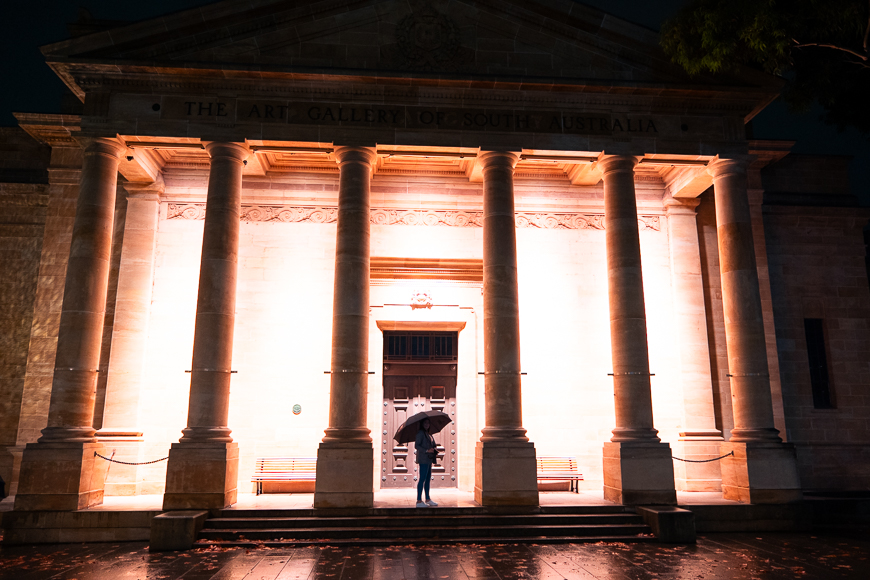 A person with an umbrella stands in front of the illuminated entrance of the Art Gallery of South Australia at night.