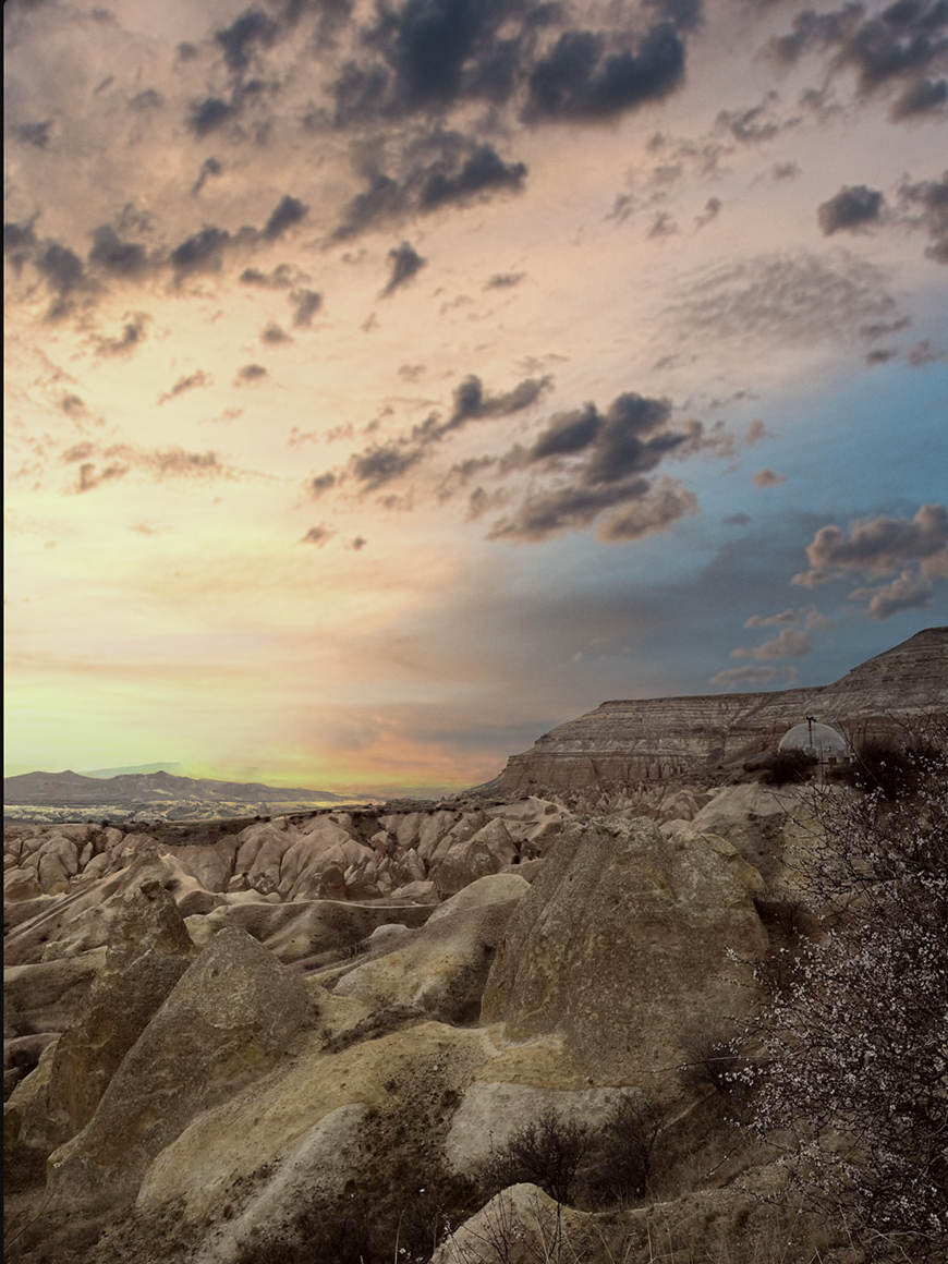 Rocky terrain with scattered shrubs under a dramatic sky at sunset, with clouds and distant mountains in the background.