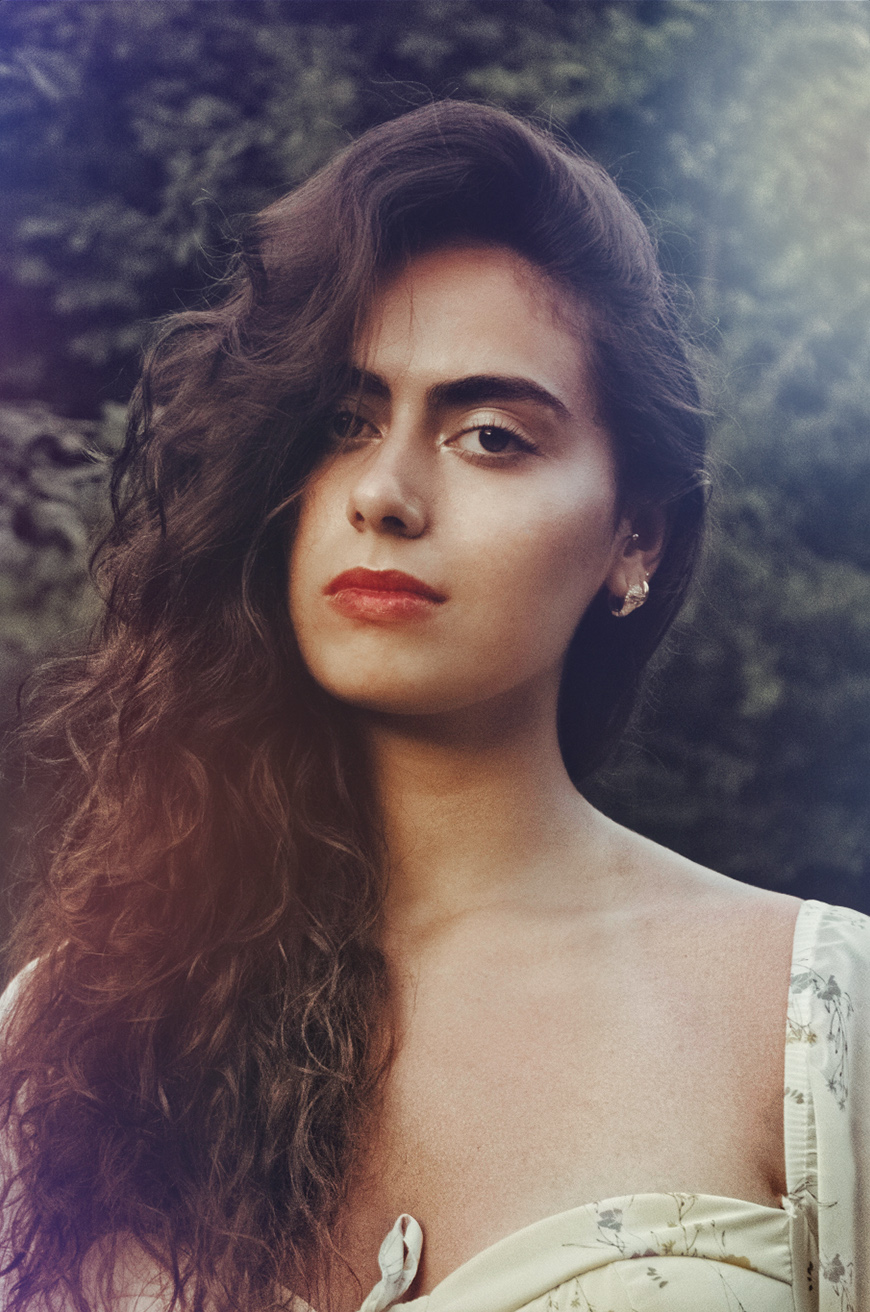 Woman with long, curly dark hair and red lipstick standing outdoors, wearing a light-colored off-shoulder top, looking at the camera with a neutral expression. Trees are blurred in the background.