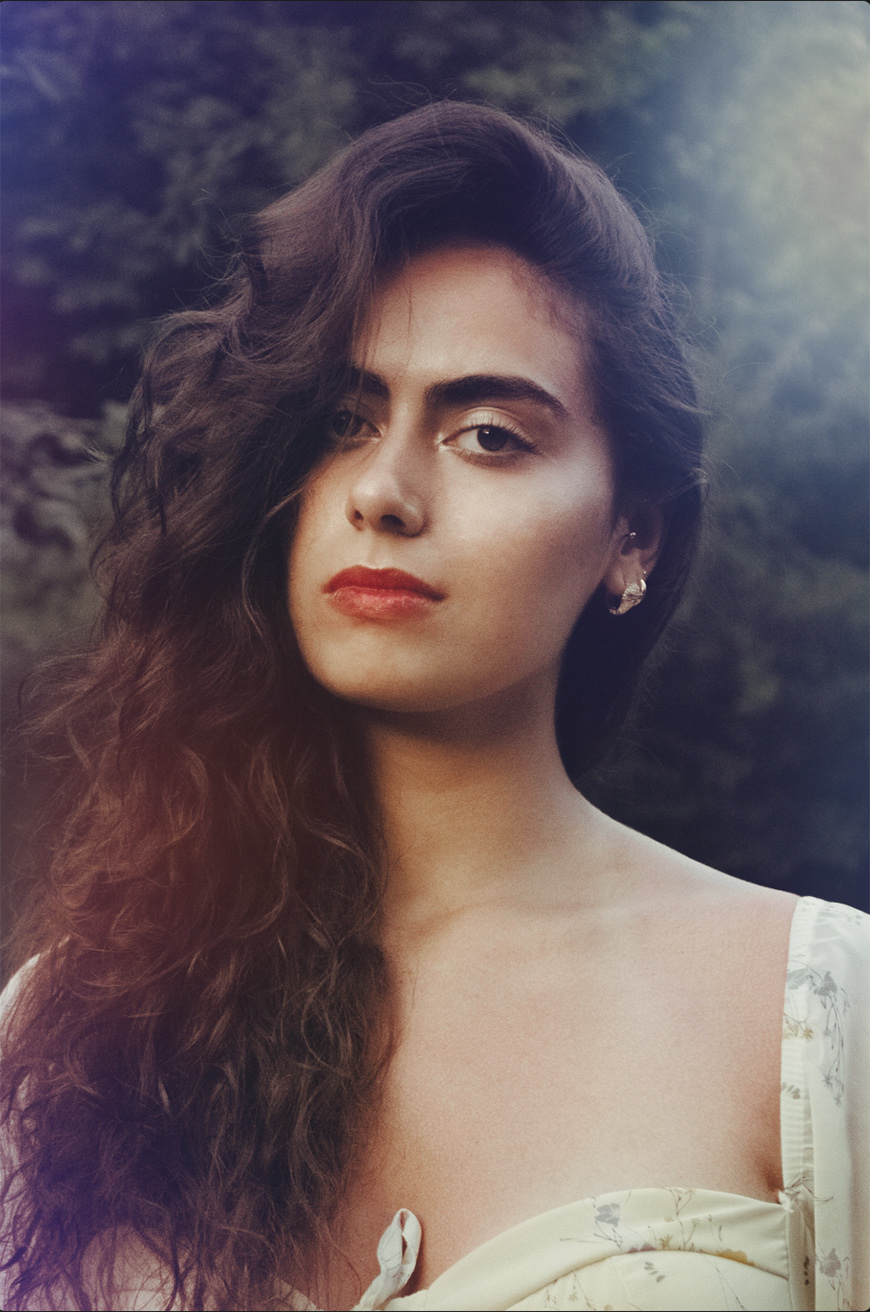Woman with long, wavy dark hair and red lipstick stands outdoors wearing a light-colored, off-shoulder top. Trees and greenery are blurred in the background.