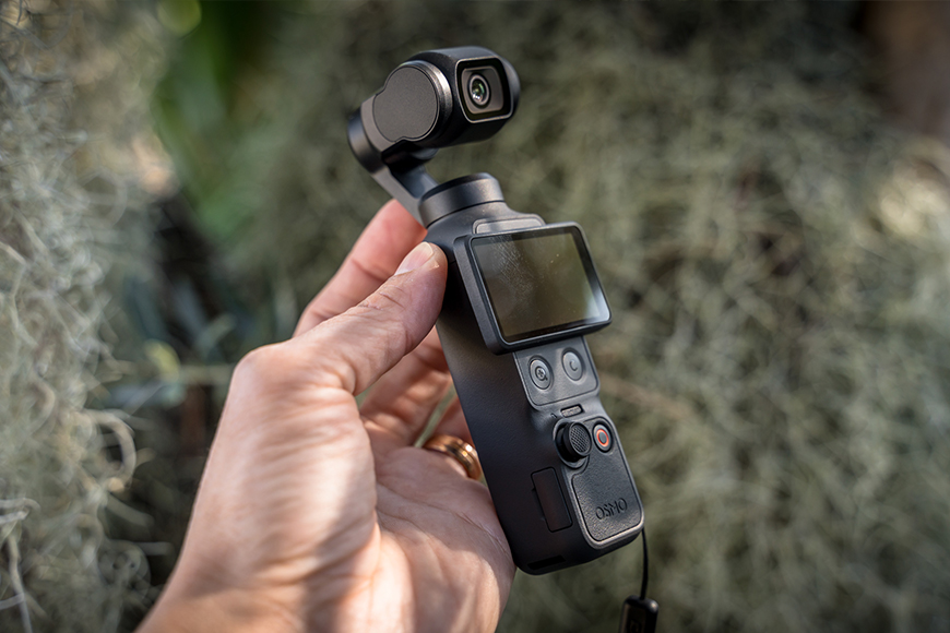 A hand holds a compact handheld camera with a small screen and control buttons, set against a blurred outdoor background.