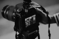 A camera on a tripod is being adjusted by a person; its screen displays a preview image of a group of people standing together.