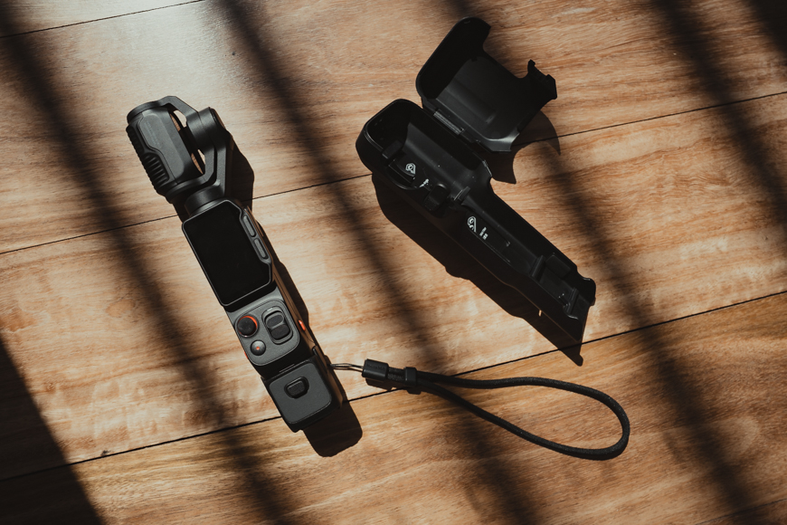 A handheld camera with a screen and wrist strap lies on a wooden surface next to its open black protective case. Diagonal shadows fall across the scene.