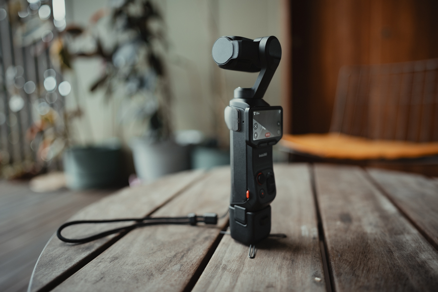 A handheld video camera with a gimbal sits upright on a wooden table indoors, with plants and a chair blurred in the background.