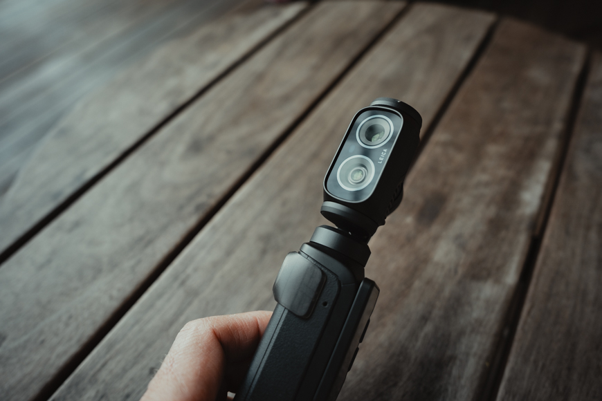 A hand holding a small black handheld camera with dual lenses, positioned above a wooden surface.