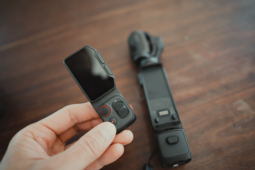 A hand holds a small black handheld camera with a screen; another similar device lies on a wooden surface in the background.